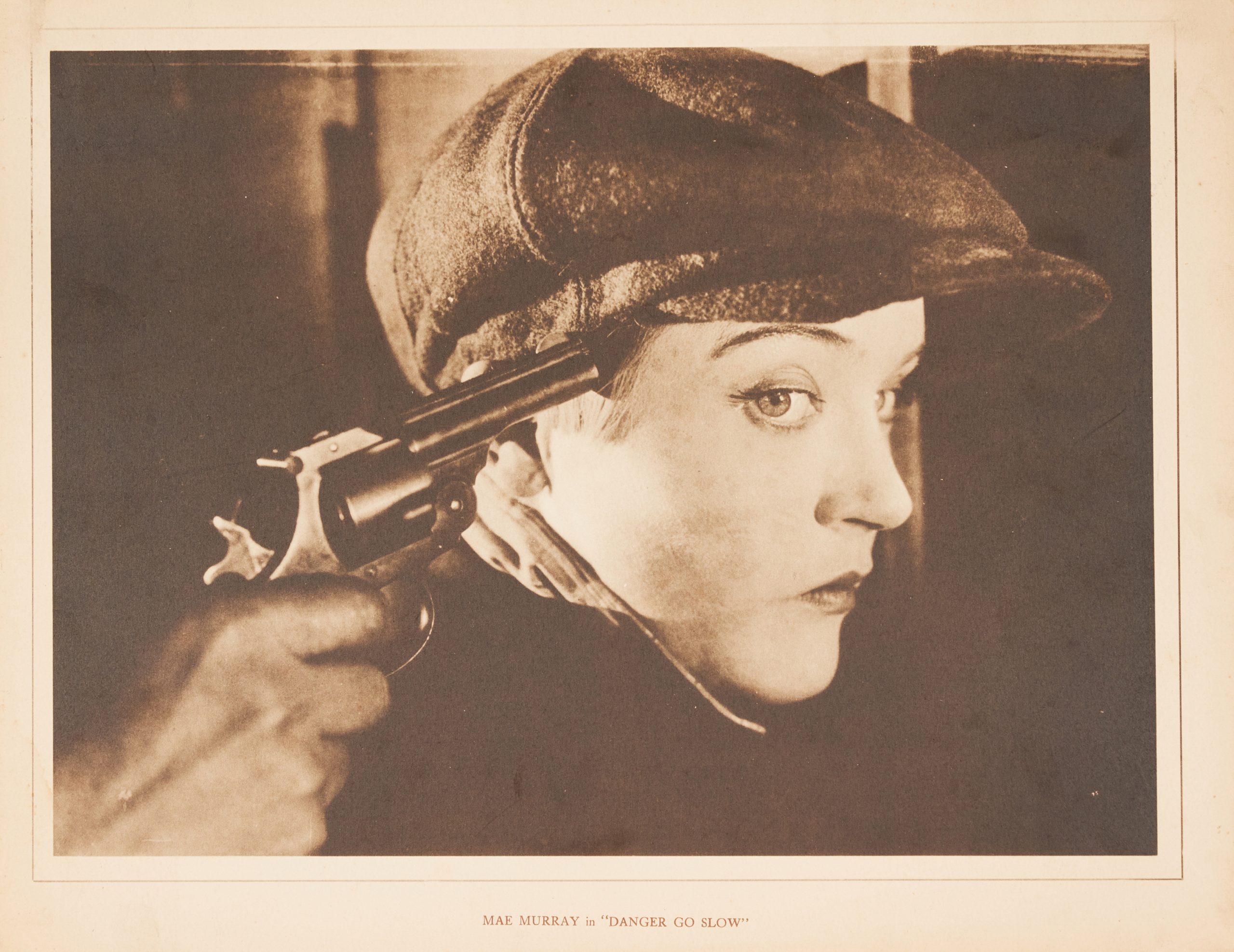A sepia-toned lobby card showing a close-up of a woman in a cap with a gun being held to her head.