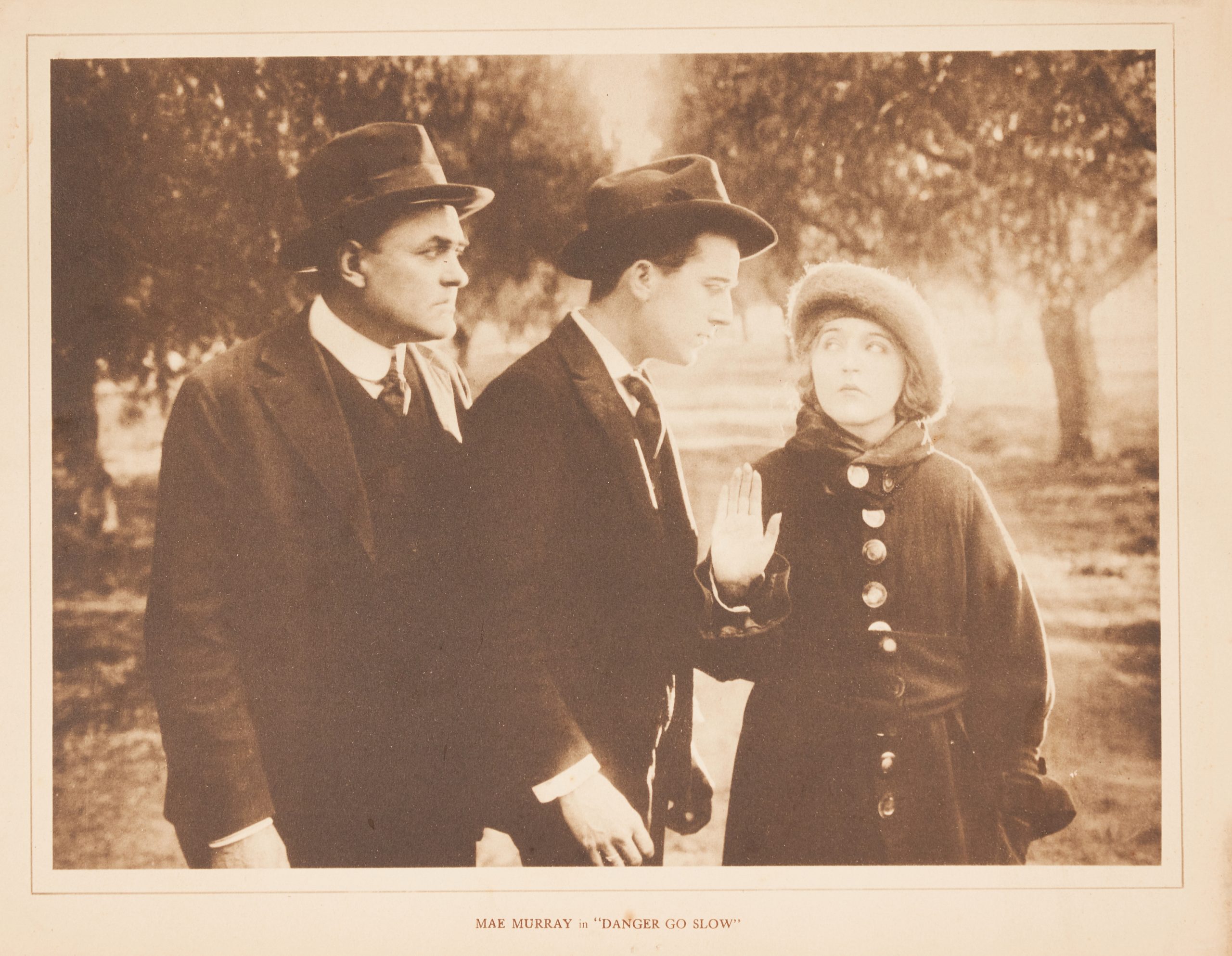 A sepia-toned lobby card with two men facing a woman with her hand up in an outdoor scene.
