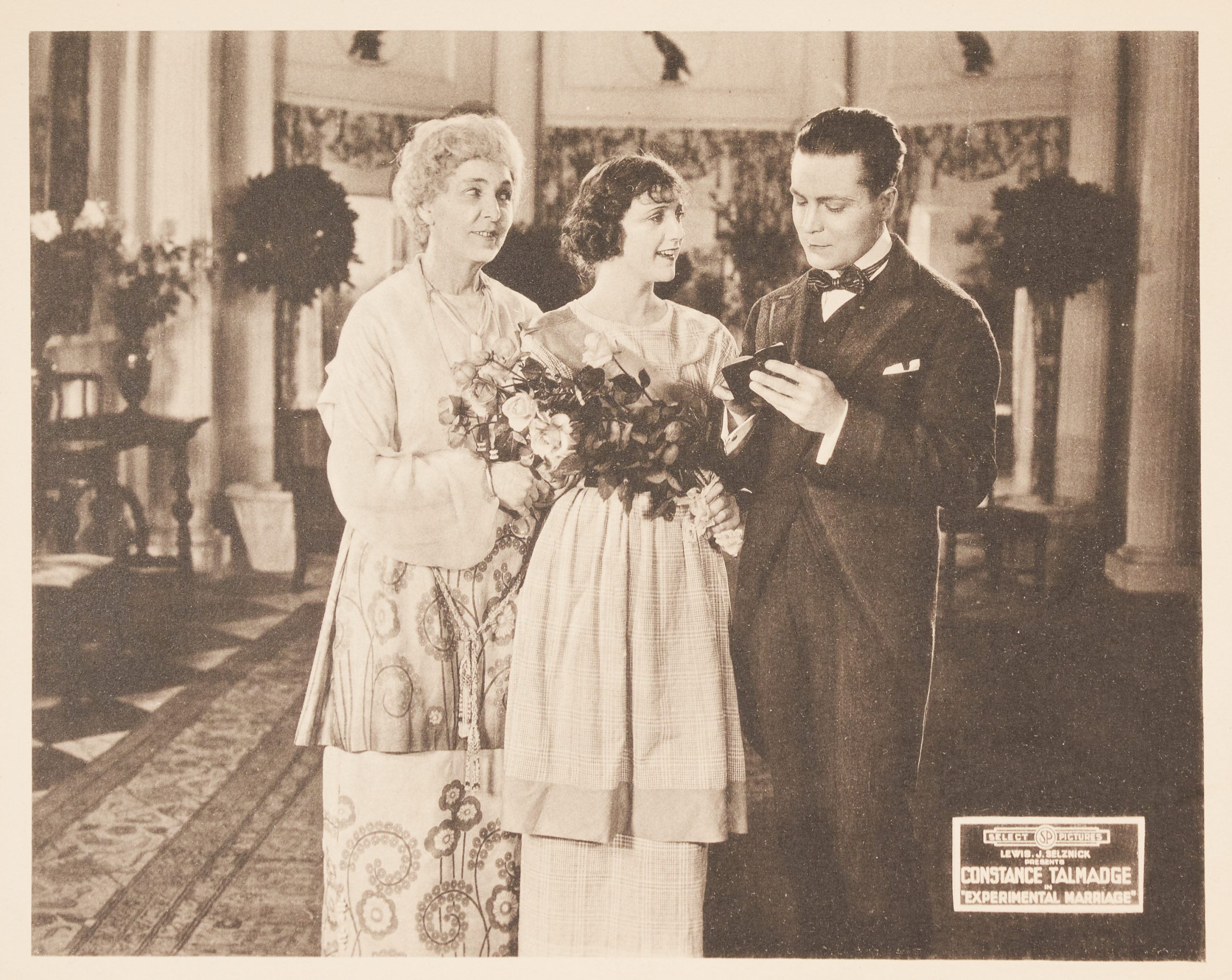 A lobby card featuring a man and two women standing in a parlor room. One woman holds a bouquet.