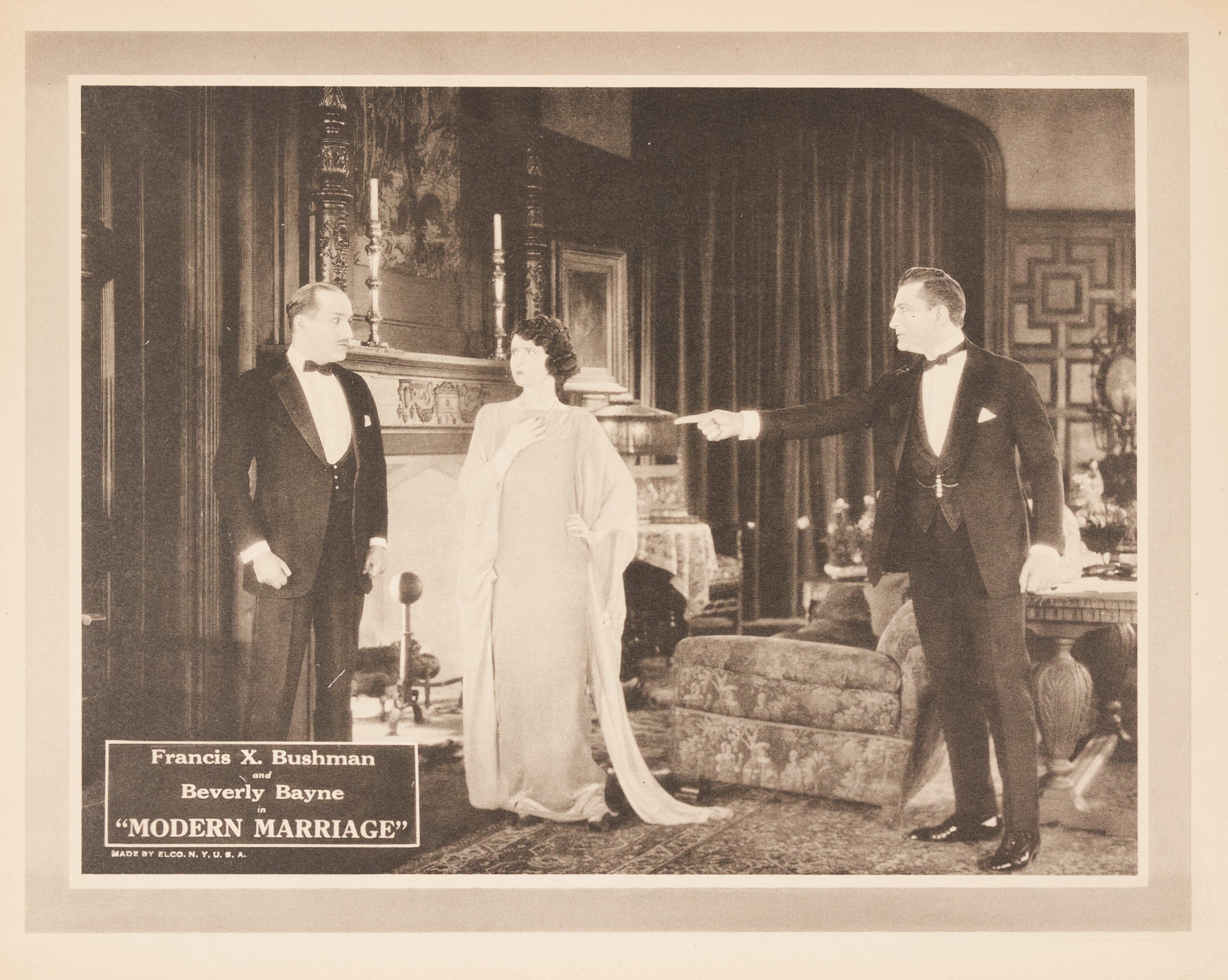 A lobby card with a woman and two men in a parlour. One man points his finger at the other man.