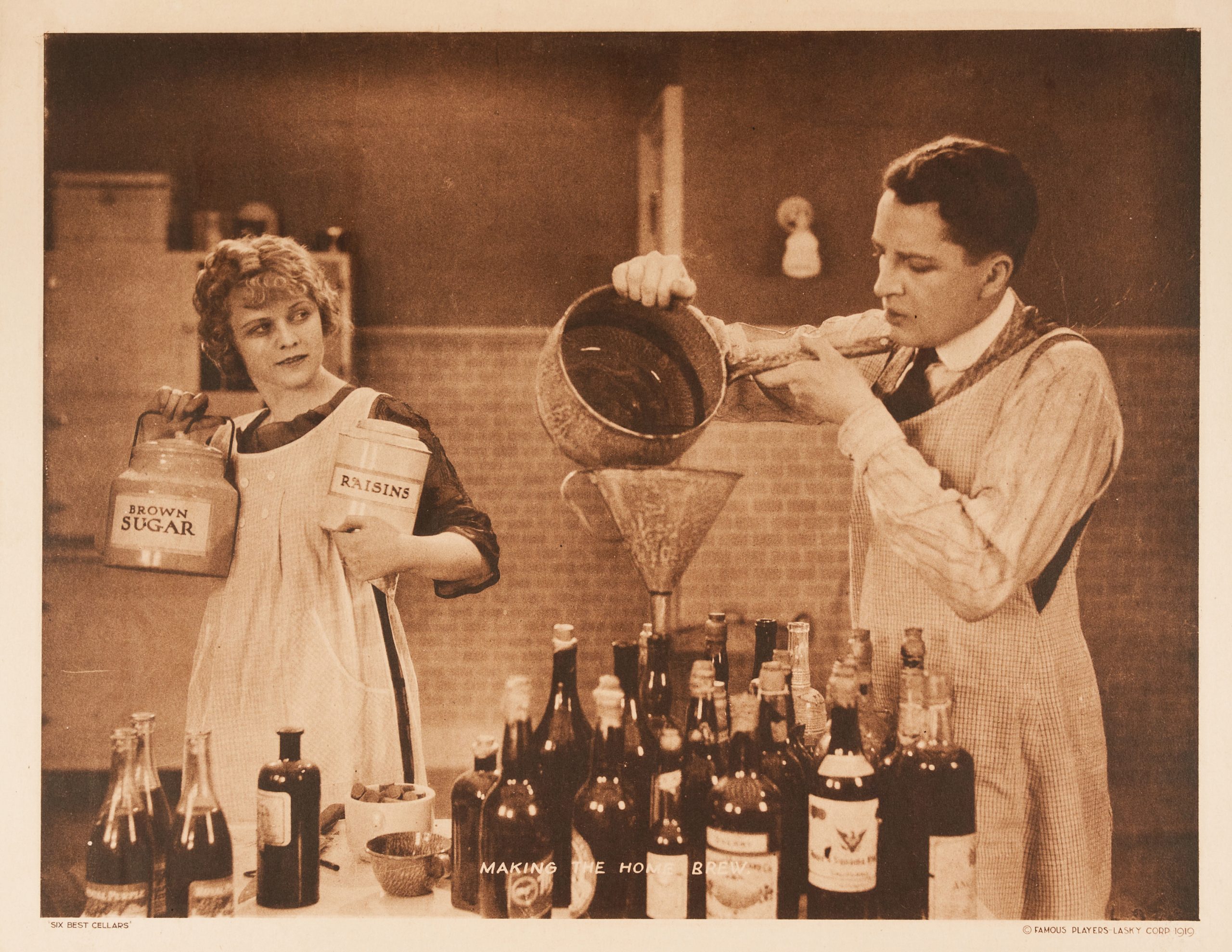 A sepia lobby card of a couple in a wine cellar. The man pours liquid into a funnel amid several bottles.
