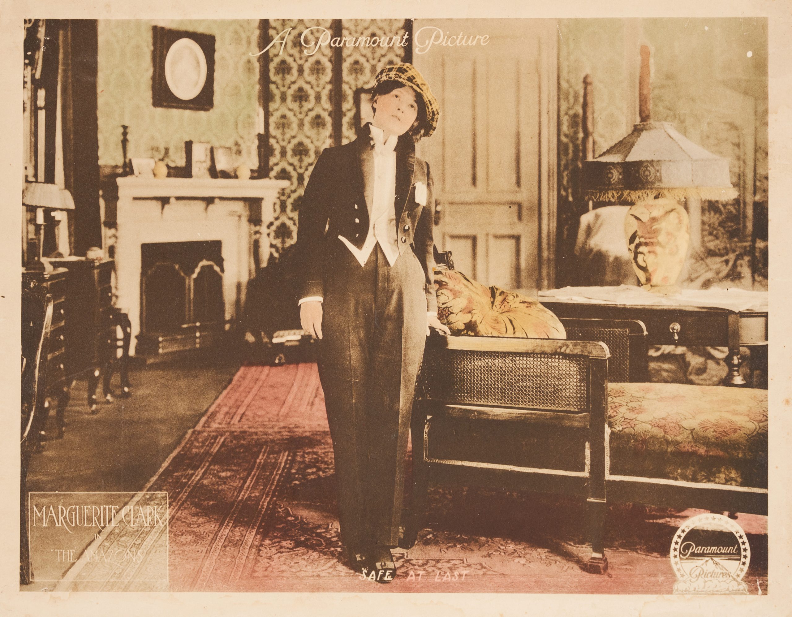 Colored-in lobby card showing a woman in pants and tailcoat standing in a well-furnished living room.