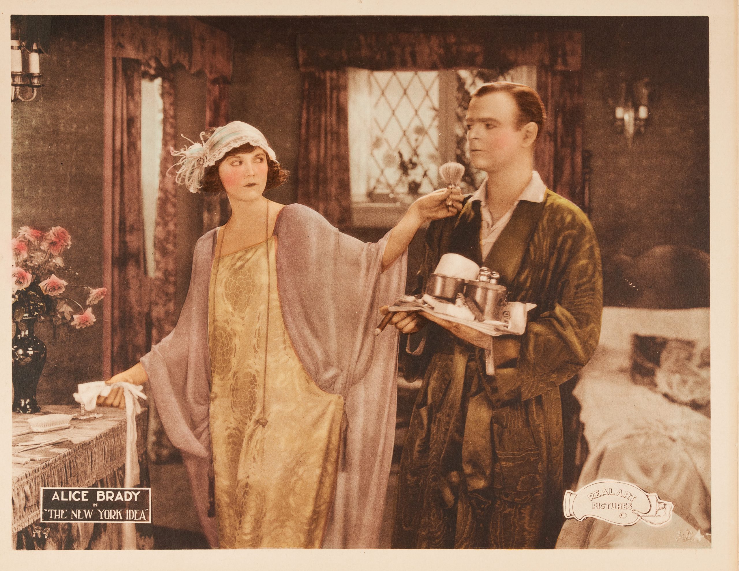 A colored-in lobby card showing a man and woman in a bedroom. She holds a makeup brush to his chin.