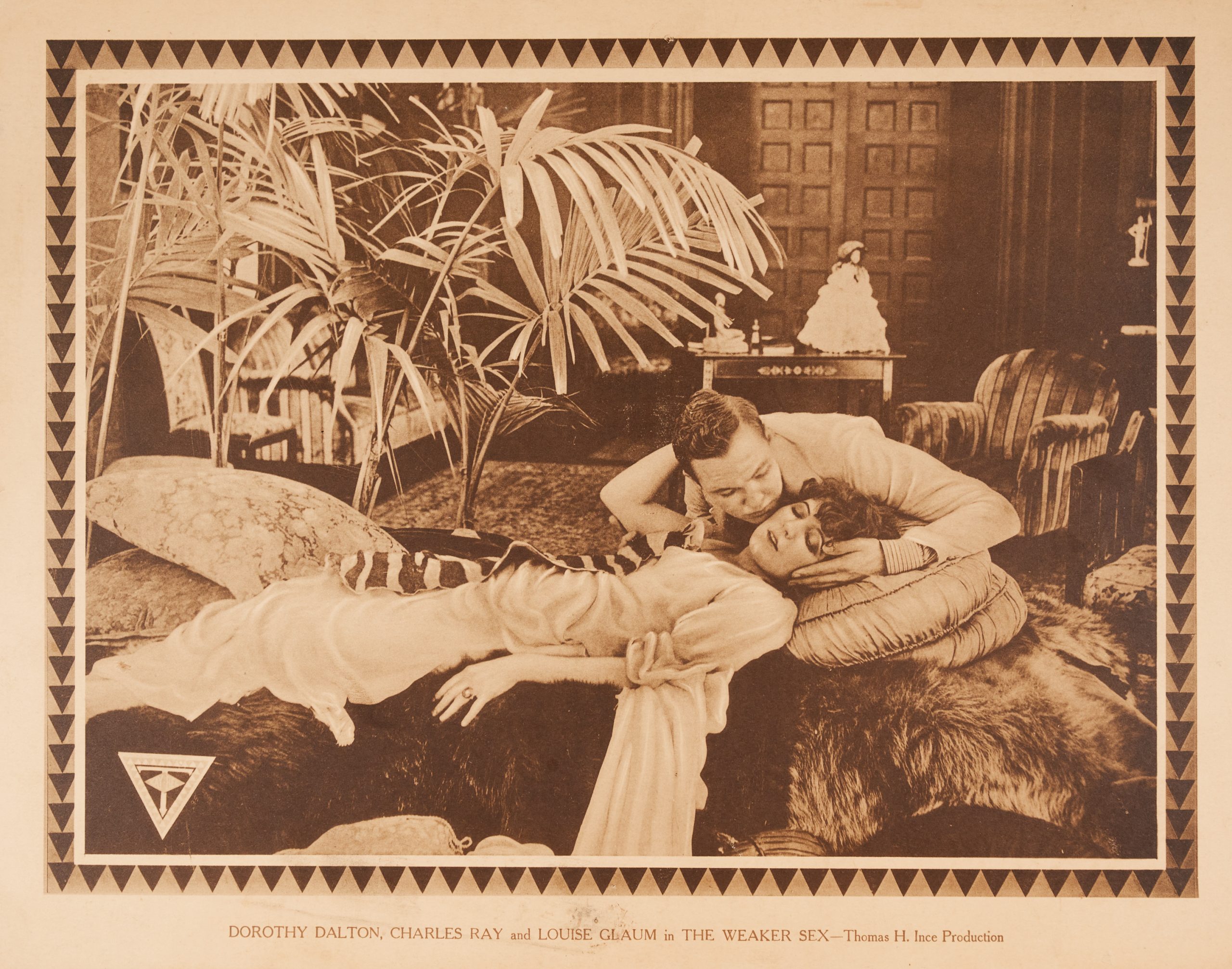 A sepia-toned lobby card with a woman lying on pillows and fur pelts while a man tries to kiss her.
