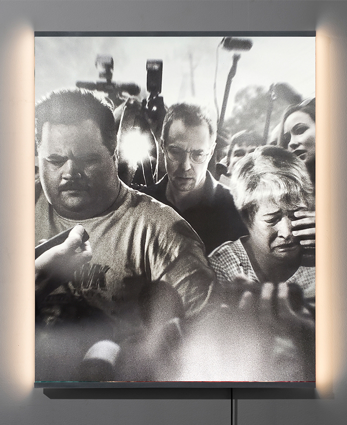 A mixed-media lit artwork depicting three distressed people walking through a crowd of cameras.