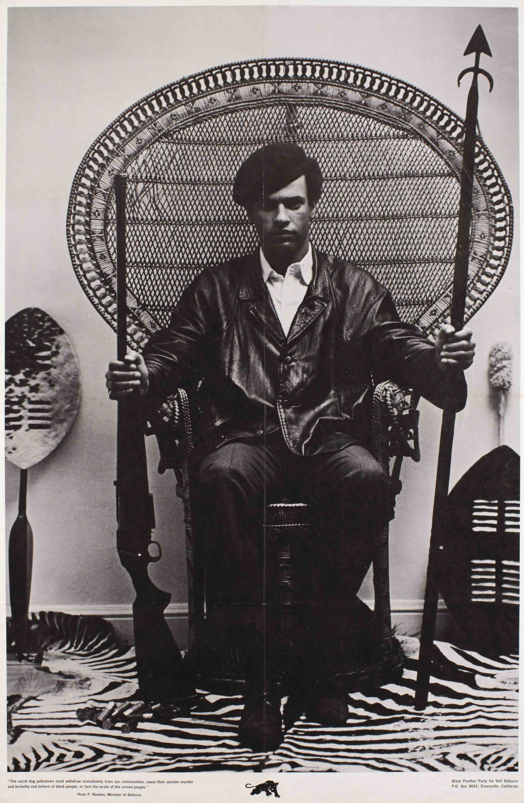A photograph of a Black man sitting on a throne-like chair holding a rifle and a spear.
